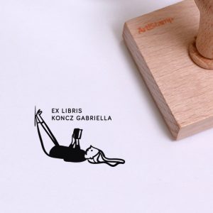 Ex libris pecsét, hanyatt fekvő, lábát falnak támasztó és olvasó lányt ábrázoló grafikával. Nyeles fa alapra készült, gumi lenyomattal, egyedileg megadott névvel készült.